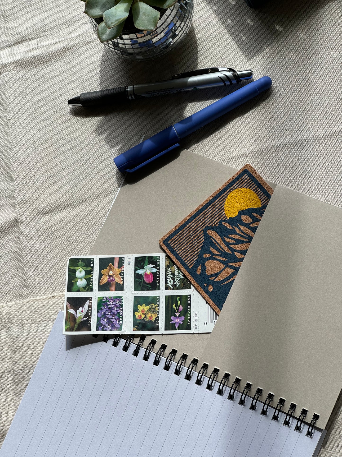 Open Spiral Notebook with an inside pocket that has stamps and a picture sticking out of it, placed on a work surface with pens and a small plant to show versatility in every day use